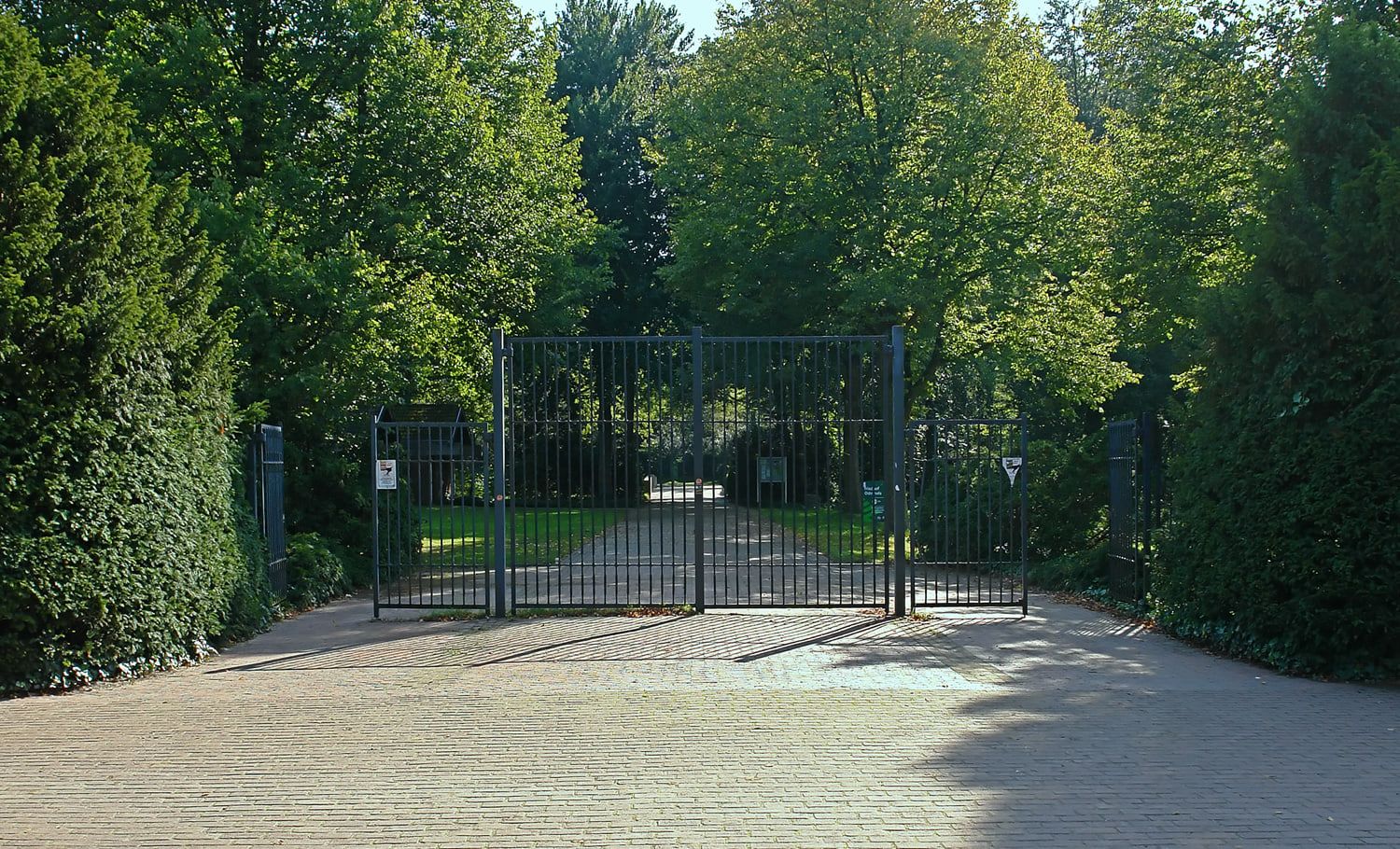 Geschlossenes hohes Metalltor an einer sonnigen, dicht begrünten Zufahrt auf Osterholzer Friedhof