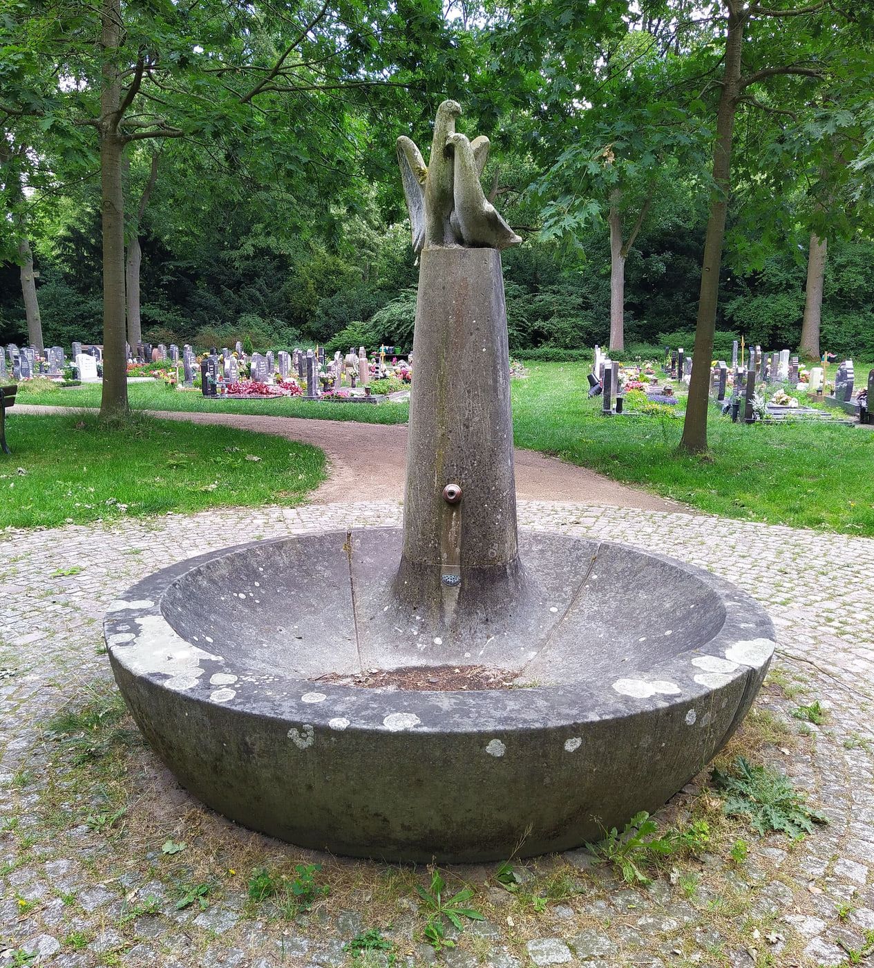 Steinerner Rundbrunnen mit Vogelplastiken vor begrünten Grabreihen auf Osterholzer Friedhof