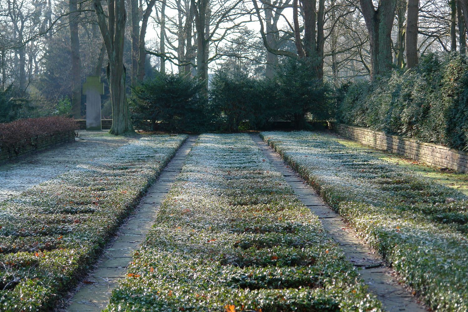 Niedrige rechteckige Pflanzfelder mit schmalen Wegen und einem Denkmal unter alten Bäumen auf Osterholzer Friedhof
